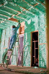 Male builder insulating wooden frame house. Man worker spraying polyurethane foam inside of future cottage, standing on ladder, using plural component gun. Construction and insulation concept.