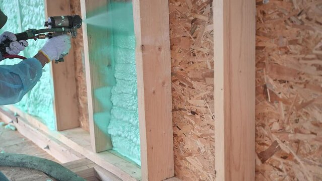 Male builder insulating wooden frame house. Man worker spraying polyurethane foam inside of future cottage, using plural component gun. Construction and insulation concept.