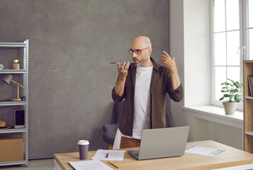 Serious professional business man on speakerphone. Bald middle aged entrepreneur standing by working desk table with notebook PC in home office, holding phone, recording thought, idea or voice message