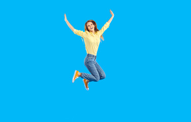 Fototapeta na wymiar Excited millennial girl isolated on blue studio background jump high raise hands celebrate success. Overjoyed young woman feel euphoric triumph with win or victory. Energetic active teenager move.