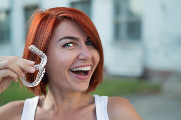 Red-haired Caucasian woman holding transparent mouthguards for bite correction outdoors. A girl...