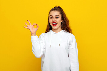 Positive good-looking young brunette woman in white casual style sweatshirt, smiling happy and doing ok sign with hand. Indoor studio shot on yellow background