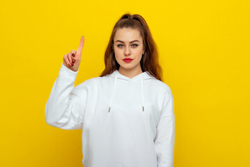 Serious beautiful brunette girl in white casual style sweatshirt, frowning, pointing fingers up, standing over yellow background