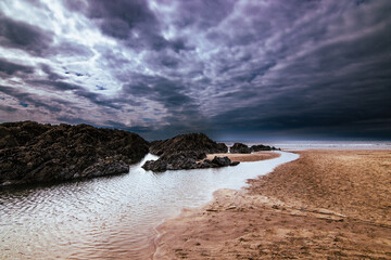 A North Devon beach between Mortehoe and Woolacombe.  Rocks, water, sand and clouds combine to bring drama.  The sun just peeks through the clouds.