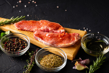 Raw meat on a cutting board. Dark background. Preparation for cooking pork meat. Various spices, seasonings lie nearby.