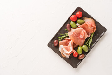 Top view on black charcuterie board with proscuitto served with olives on white concrete table...