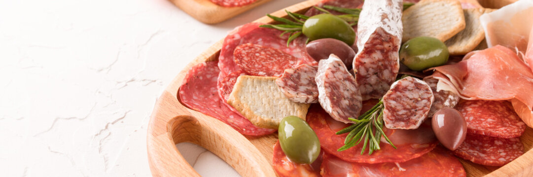 Banner with assortment of different meat snacks, charcuterie wooden board with different types of sausages. Gray plate with traditional italian antipasti on white table with copy space