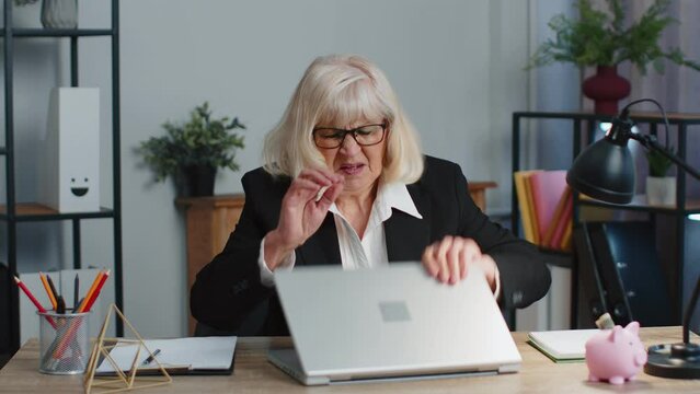 Irritated senior mature business woman angrily hits table with fist, closes laptop. Broken laggy buggy computer pc. Remote working, social distance elearning, training, online webinar at home office.