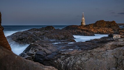 Attractive night seascape, sea waves splashing over rocks, shining lighthouse. Evening darkness, long exposure, blurry water trails and misty steam. Southern Black Sea coast, Ahtopol, Bulgaria.