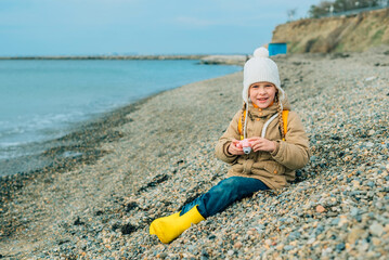 Fototapeta na wymiar little girl sits in autumn, spring on the beach, looks at the camera with a smile, holds a children's pink camera in her hands. the child is passionate about his hobby: photography