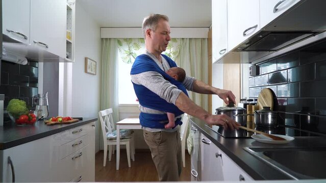 Father with little baby sleeping in sling cooking in the kitchen. Man on paternity leave working household chores. Dad enjoying fatherhood responsibility caring for child