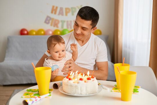 Portrait of father with adorable toddler baby celebrating first birthday, cake is surprise for child, make a first wish, posing at home with balloons and party inscription on background.