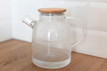 Glass teapot with water standing on wooden countertop against white wall background.