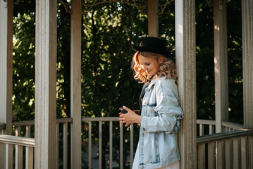 Smiling young woman with curly hair and cup of coffee in park on sunny evening. Cute hipster model in hat and denim jacket enjoying evening in wooden rotunda, outdoors