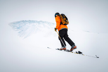 Winter skitour freeride in cloudy weather, snow-capped mountains against the backdrop of a glacier. Skier man in full gear climbs uphill in a skitour