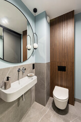 Modern bright bathroom with white toilet seat on wooden lamela wall. Round mirror with black frame.