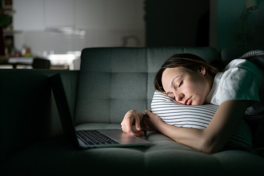 Exhausted young woman sleeping on couch after working with laptop late at night at home. Tired freelancer girl fell asleep taking a nap lying with computer. Overworking, tiredness concept