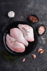 Chicken breast. Raw chicken breast fillets on black ceramic plate on wooden cutting board with herbs and spices on black background. Top view with copy space.