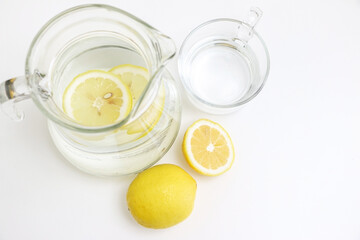 Top view of refreshing and healthy natural lemon water, lemonade, in a glass jug, and glass cup, besides full and sliced lemon, on clean white table