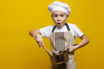Isolated photo of a surprised European teenage boy in an apron and a chef's hat, holding a saucepan and a wooden spoon in his hands, tasting, cooking a surprise dinner for his parents.