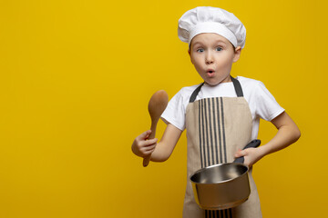 Isolated photo of a surprised European teenage boy in an apron and a chef's hat, holding a saucepan and a wooden spoon in his hands, tasting, cooking a surprise dinner for his parents.