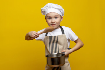 Funny little chef in a chef's hat and apron is cooking something in the studio, holding a saucepan and a wooden spoon in his hands, showing surprise against an isolated background.
