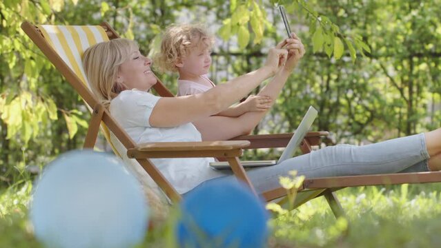 Happy Smiling Family In Green Garden, Sunny Summer Day. Mom Uses Cell Phone Take A Selfie With Her Little Girl Child. Together Laughing To Smart Phone Camera. Mother With Daughter Surfing Internet.