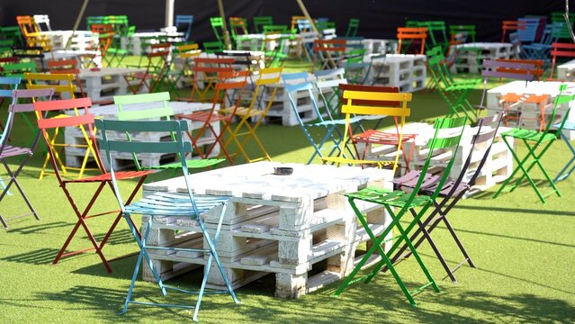 dehor outdoor space with white wooden tables produced with pallets for shipping and colorful chairs in many colors with green synthetic grass carpet, outdoor setting of pub on the Navigli in Milan