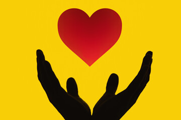 Obraz premium The silhouettes of two hands, below as a cup (open palms), receiving a drawn red heart shape. Black shapes on a yellow background. 