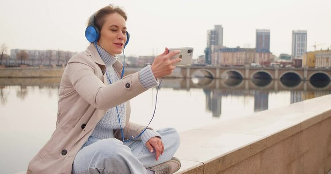 35 Year Old Woman During An Online Video Call. She Sits On The Embankment And Communicates With The Interlocutor Via Video Communication.