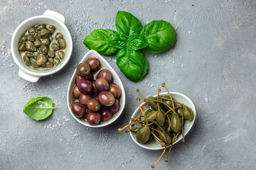 Caper berries and olives on white plate with fresh basil leaves on a gray background. Mediterranean snack assortment. banner, menu, recipe place for text, top view