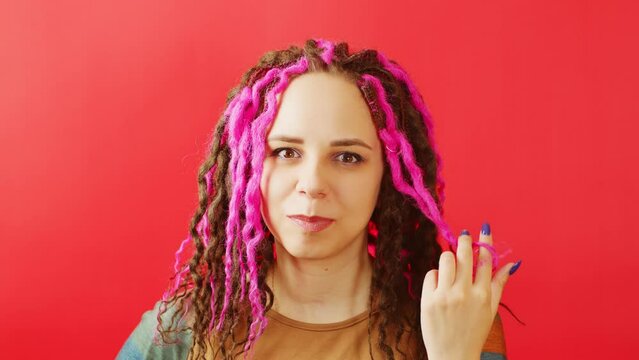 Young woman twisting hair and looking at camera on red background. Portrait of pretty female with curly hairstyle, dreadlocks posing in studio.