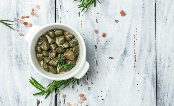 Marinated capers with fresh rosemary in a small bowl, on light wooden background. Long banner format. top view