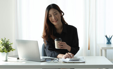 Smiling Asian businesswoman holding a coffee mug and laptop at the office. Looking at the camera.