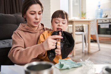 Kid with down syndrome sorting garbage near his mother at the table at home