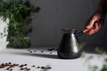 Freshly brewed aromatic coffee in cezve. A woman`s hand puts a coffee pot on a table with coffee beans and green plants.