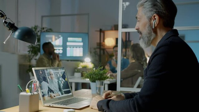 Cheerful businessman making video call talking to muslim businesswoman using laptop and earpods at night in workplace. Communication and people concept.