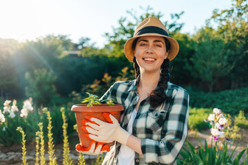 Portrait of a young smiling woman in a straw hat holding a pot of seedlings. Summer garden on the background. Earth Day concept