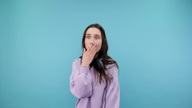 Cute lady in sweatshirt and headphones around her neck shows a gesture of silence and looks in camera, isolated on a blue background.v