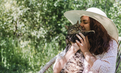 Happy young adult woman in hat sits in garden outdoors and cuddling with a cute cat.
