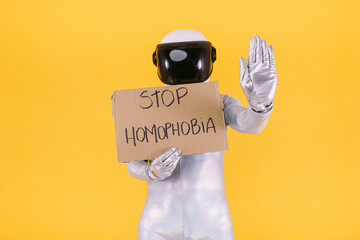Gay man dressed as an astronaut with a helmet and silver suit, holding a sign that reads: 'STOP HOMOPHOBIA' and raising his hand as a sign of stopping, on a yellow background.