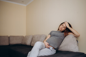 Young beautiful pregnant woman sitting at home on the couch sick, has a headache and nausea