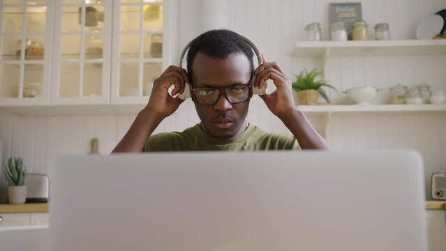 African American student puts on headphones to listen audio courses in laptop. Man in glasses sits against countertop in kitchen closeup slow motion