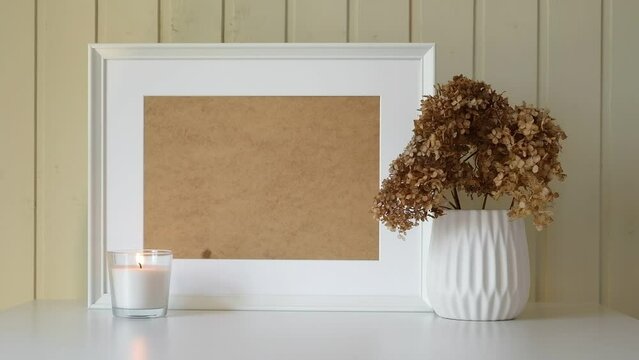 Empty picture frame with beautiful bouquet of dried hydrangea flowers. Modern white ceramic vase with dry grass. Wooden wall background. Elegant Scandinavian interior. Fall still life. Burning candle.