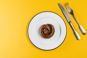 Plastic excrement lies on a plate between a fork and a knife close-up. The concept of an inept cook, fake news, shitty restaurant, bad food, taste of shit