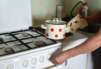 An elderly woman puts a pan on a gas stove with a hand in a mitten, turns on a burner with the other hand.