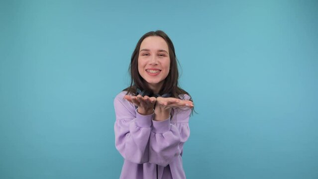 Positive girl in a sweatshirt and headphones around his neck shows a heart gesture with a smile on his face and sends an air kiss and smiles