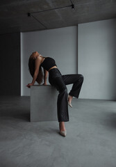 Fototapeta na wymiar teen fashion. young brunette girl in black top and leather pants sits fashion in profile on the gray cube and looks up on the white wall background in loft interior. fashion concept, free space