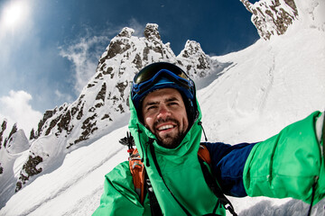 Young smiling skier in helmet, googles and ski jacket takes a selfie against sun and beautiful snow...