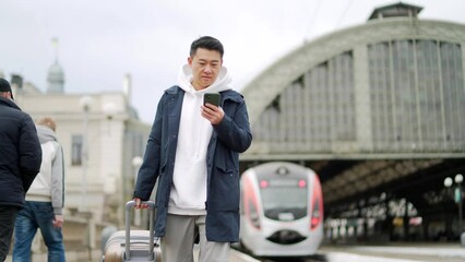 Asian tourist booking accommodation at home using mobile phone, passenger arrives by train to new city. Passengers at rairoad station and arriving train. Man use smart phone before travel